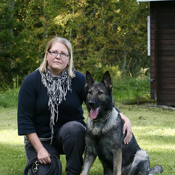 Omistaja poseeraa harmaan saksanpaimenkoiran kanssa nurmipihalla. Omistaja polvilla maassa, koira istuu vieressä kainalossa.